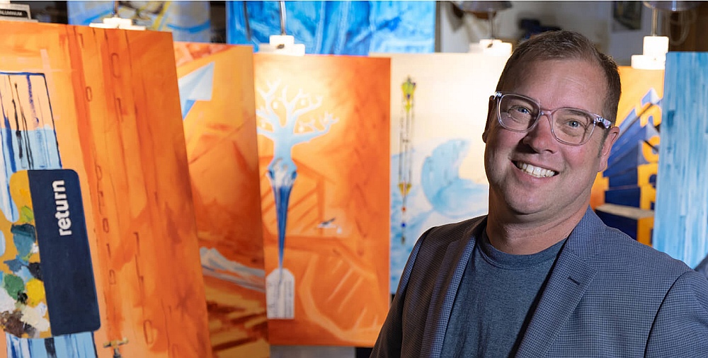Photo of Vice President and Creative Director, Jim Coe, in front of several of his own paintings at an art show at the Providence Cancer Center.