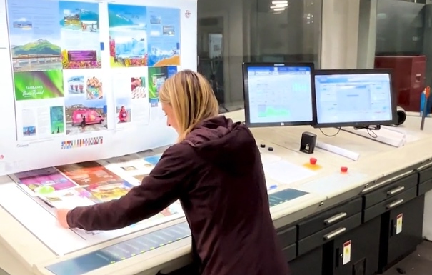 Our Creative Director, Tara Storter, examines large printed sheets on a wide table also featuring computer monitors and a vertical poster with additional graphics.
