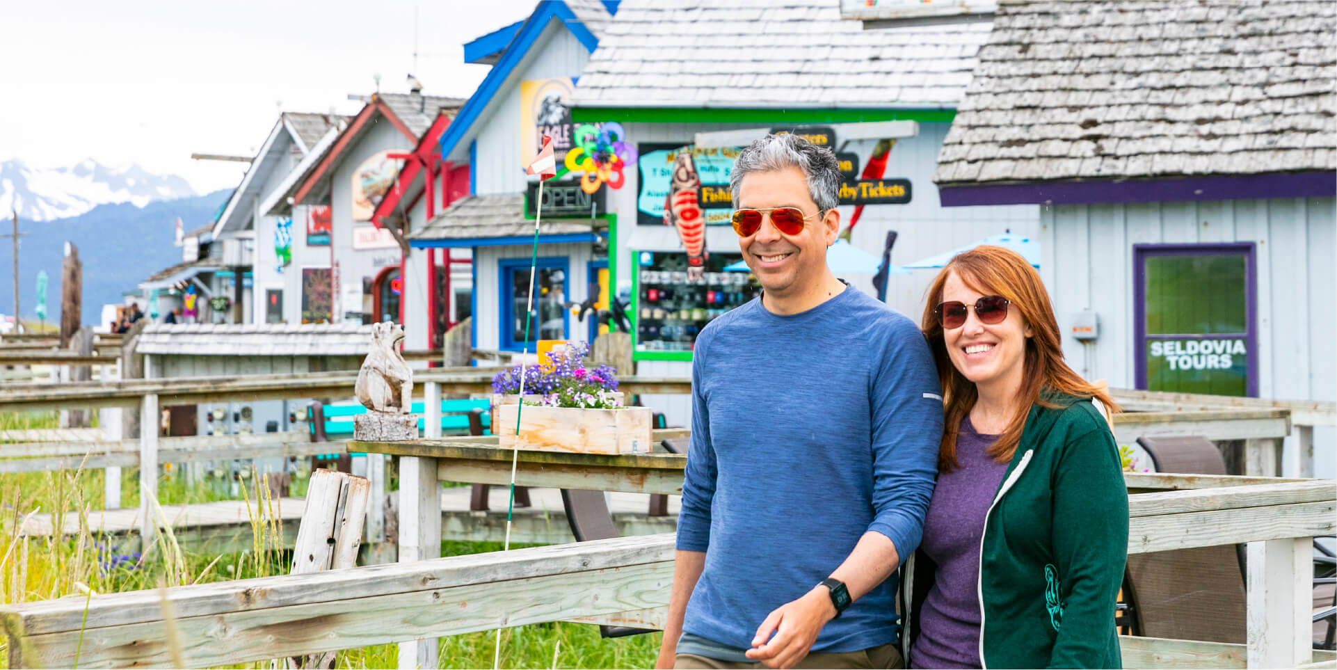 Two people walking along the wooden boardwalk in Homer, Alaska, with colorful waterfront shops, flowers, and mountains in the background.