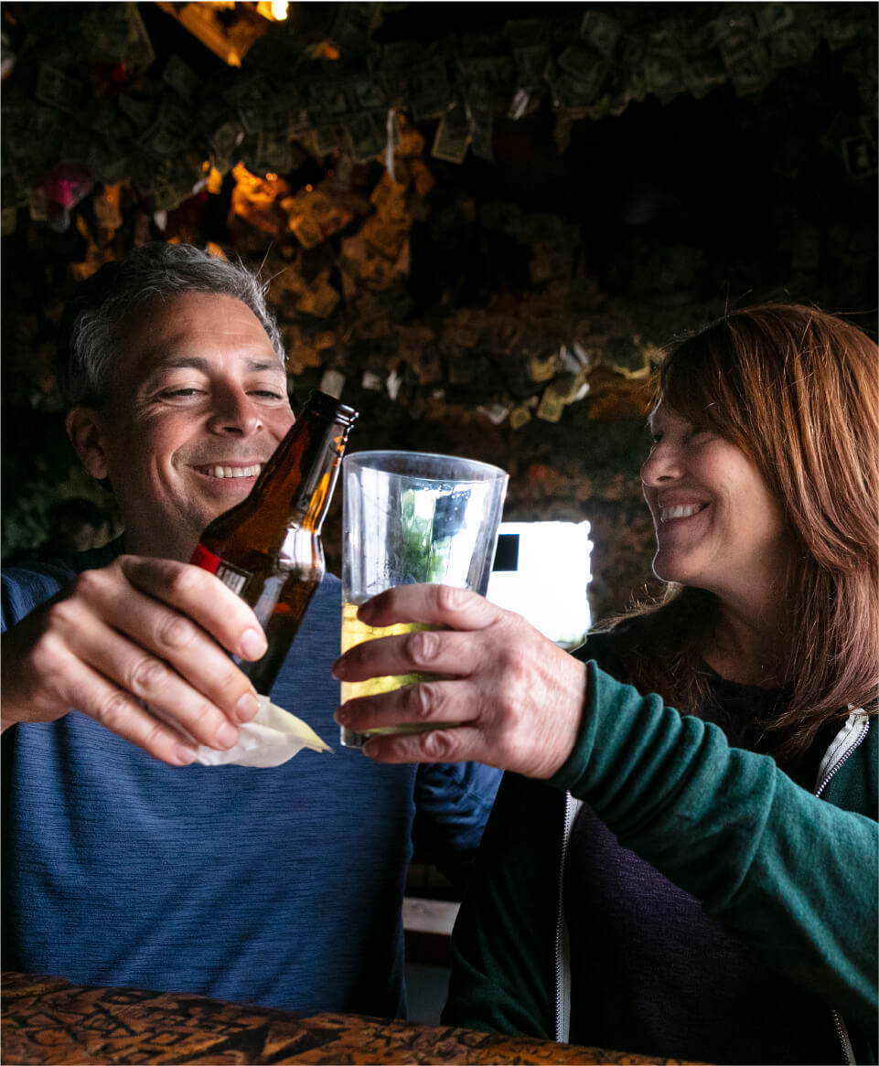 wo people clinking drinks inside the Salty Dawg Saloon, with a beer bottle and a glass raised over a bar counter and walls covered in pinned dollar bills in the background.